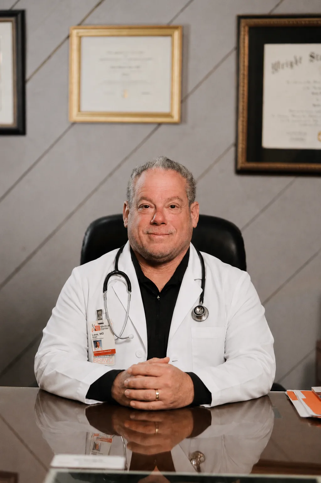 Dr. Brian Leve seated at his desk in a white coat with a stethoscope, framed diplomas on the wall behind him.
